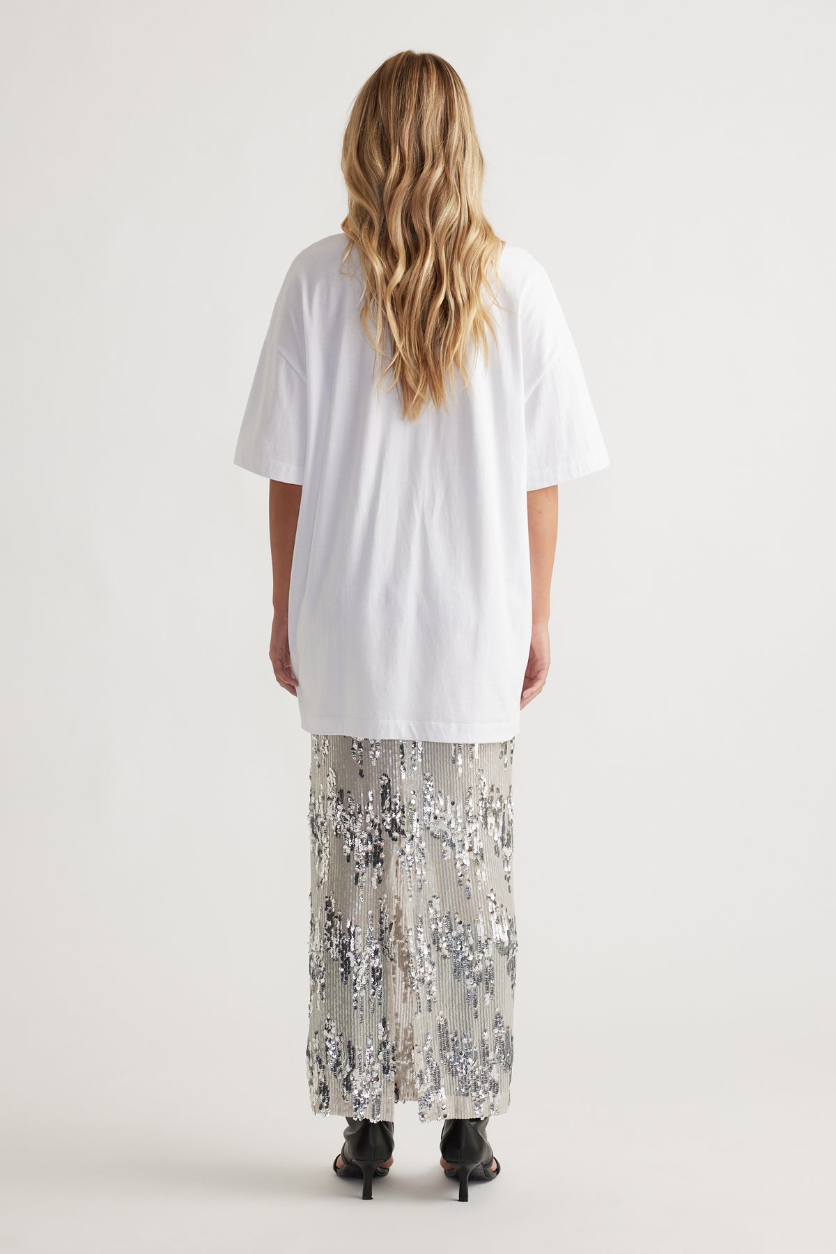 Woman seen from behind wearing a relaxed white longline tee paired with a shimmering silver sequin skirt and black heels