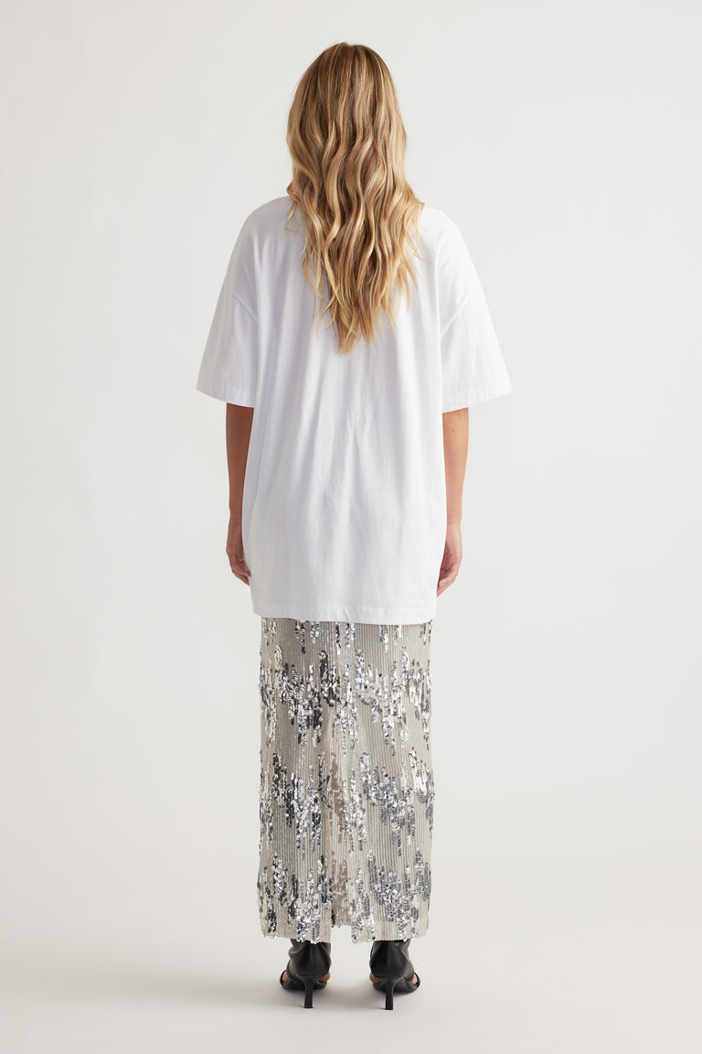 Woman seen from behind wearing a relaxed white longline tee paired with a shimmering silver sequin skirt and black heels