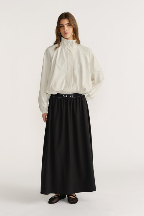 Woman wearing a high-rise black pull-on ankle-length skirt paired with a loose white zip-up jacket and flats