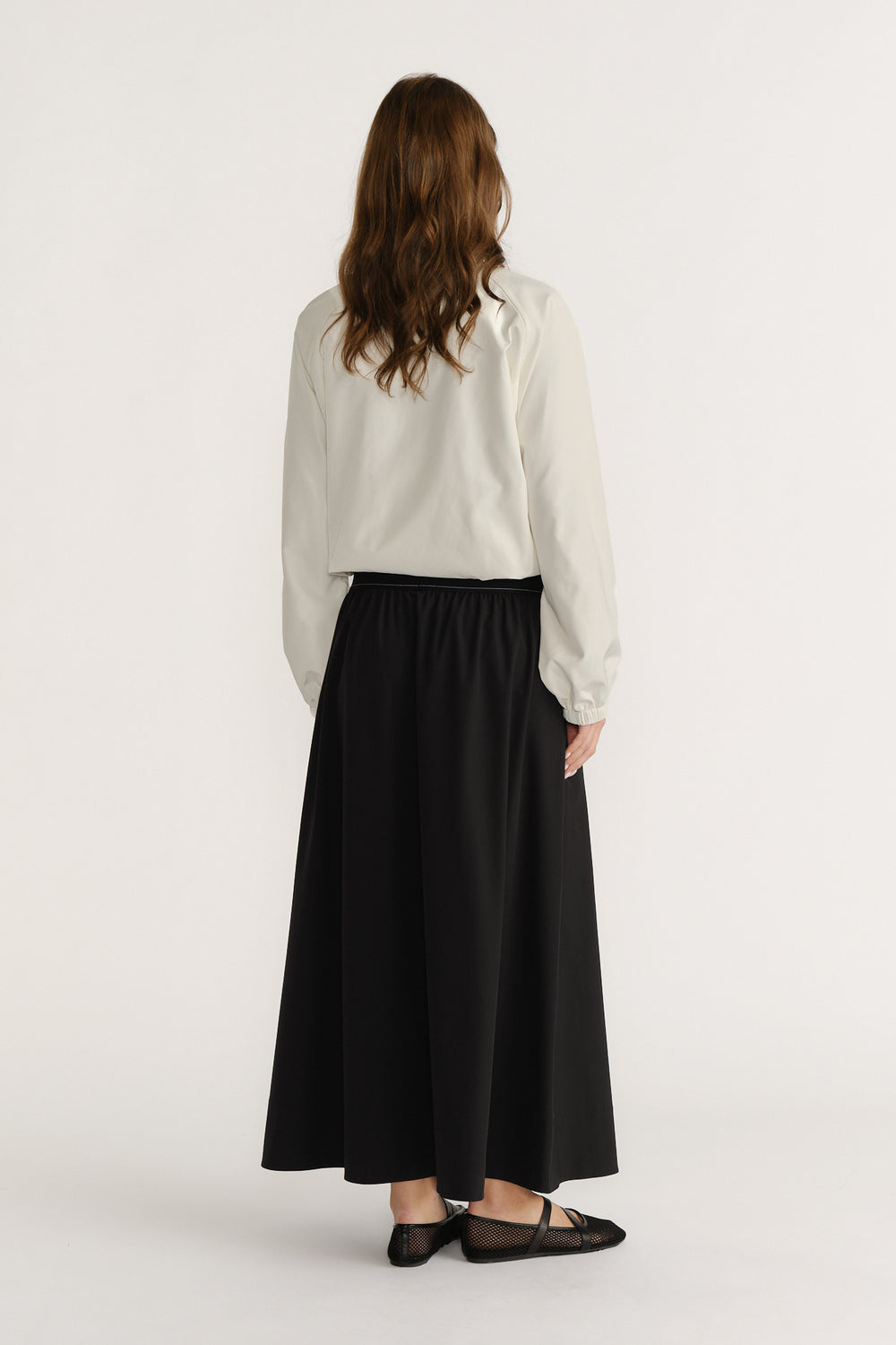 Woman wearing a black ankle-length A-line skirt with elastic waist paired with a cream long sleeve top seen from behind