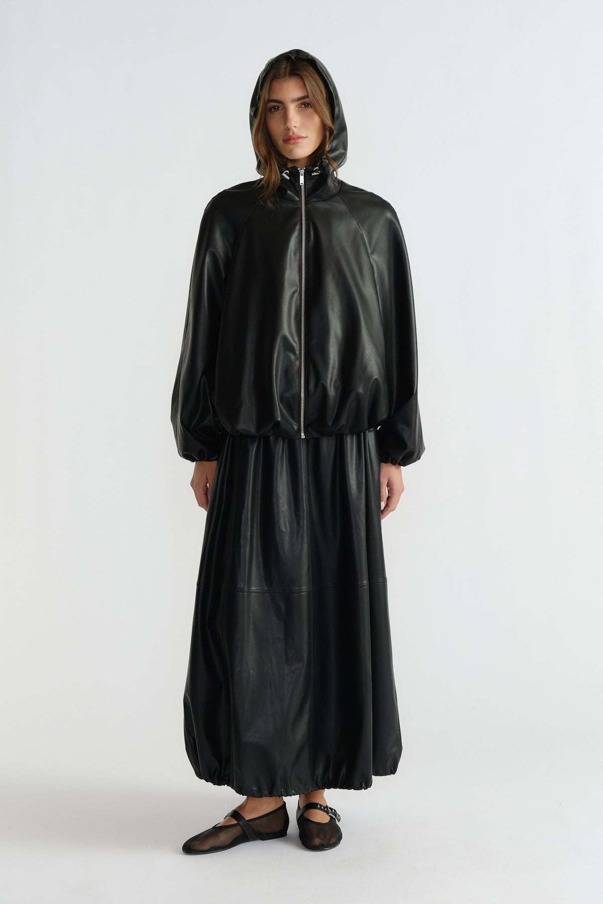 Woman wearing a sleek black faux leather bomber jacket with hood and matching long skirt, styled with sheer flats