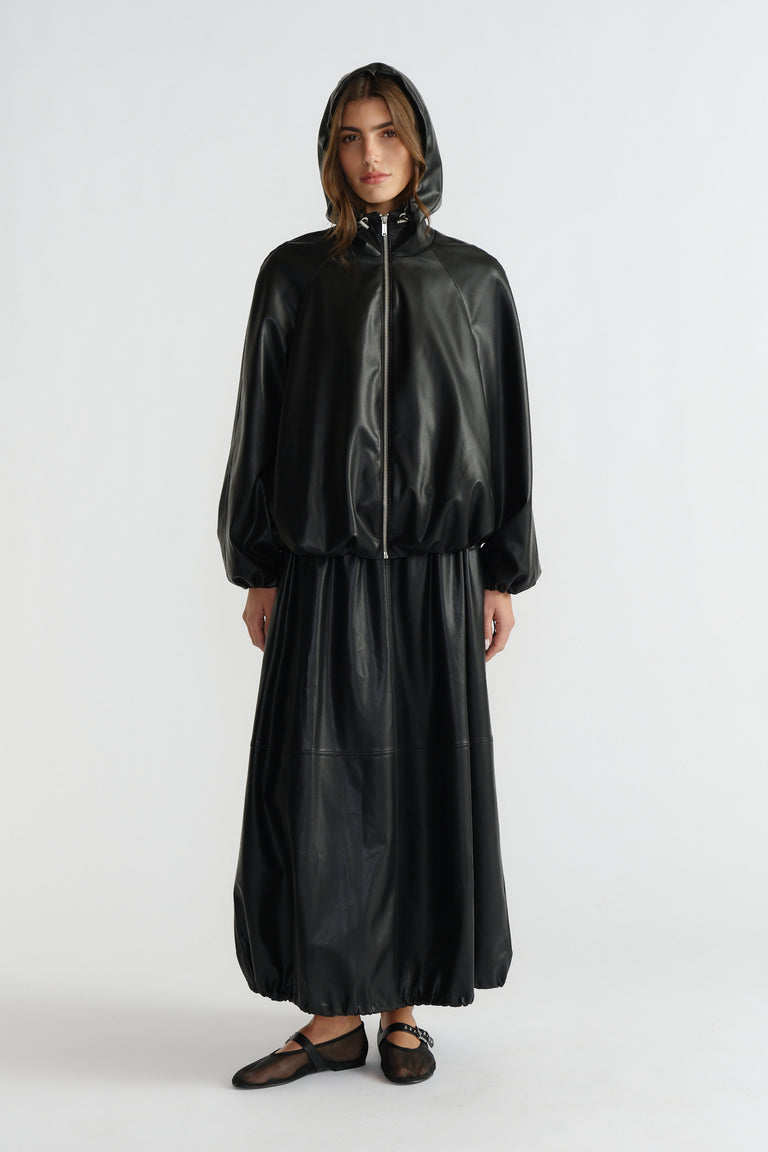 Woman wearing a sleek black faux leather bomber jacket with hood and matching long skirt, styled with sheer flats