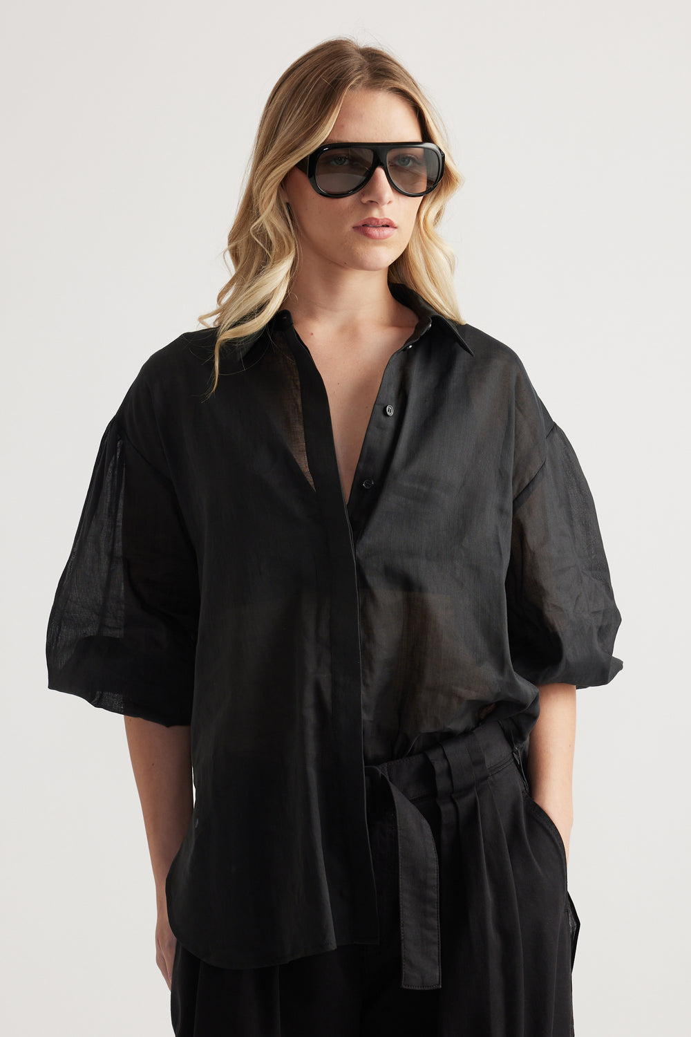 Woman wearing a sheer black oversized button through shirt with relaxed sleeves and sunglasses