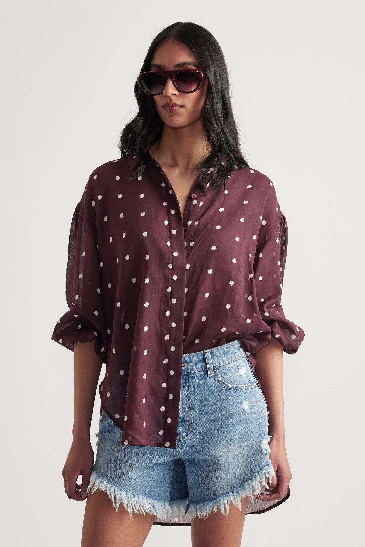Woman wearing a burgundy polka dot button through shirt with denim shorts and sunglasses
