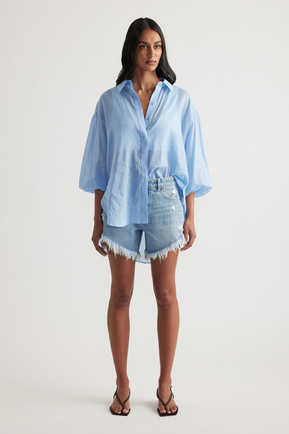 Woman wearing a light blue oversized button up shirt with denim cut off shorts and strappy sandals