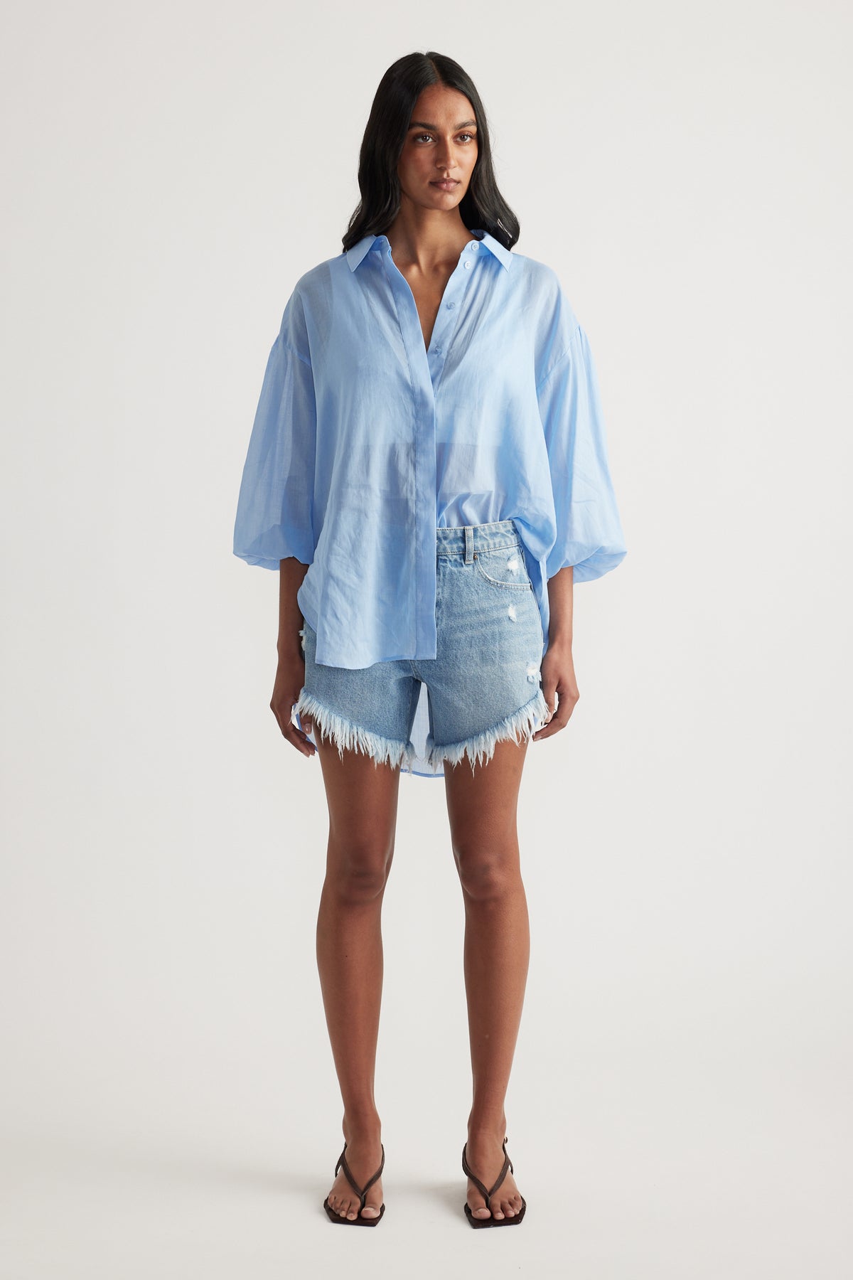 Woman wearing a light blue oversized button up shirt with denim cut off shorts and strappy sandals