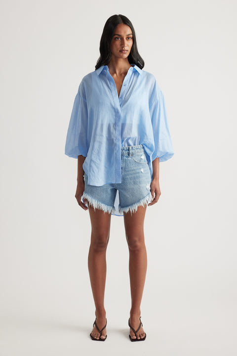 Woman wearing a light blue oversized button up shirt with denim cut off shorts and strappy sandals