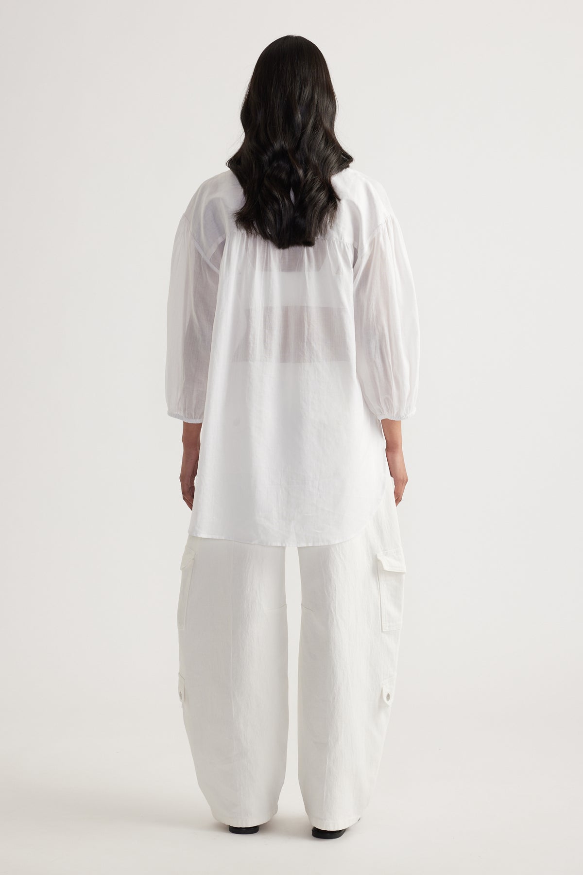 Back view of a woman in a white oversized collared shirt with long sleeves, white pants and black shoes