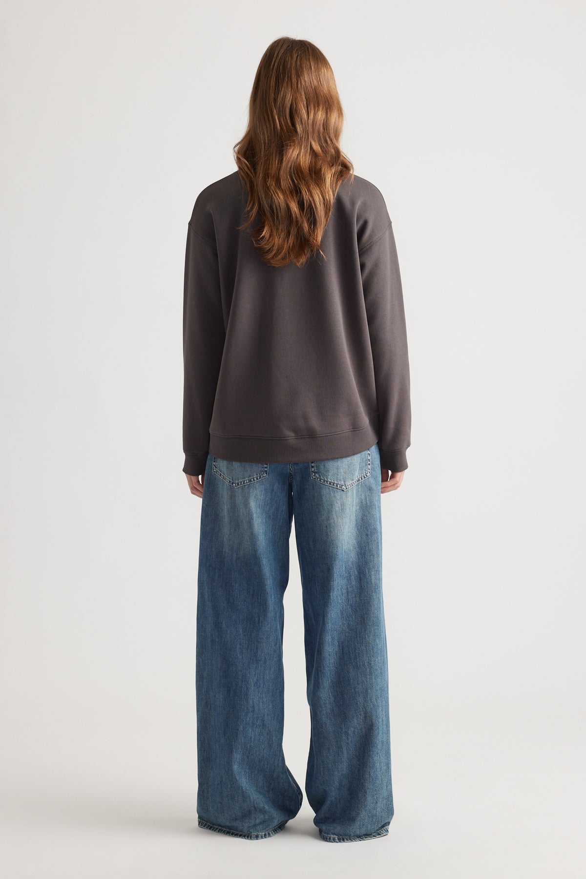 Woman wearing a relaxed dark crew neck sweat paired with wide-leg mid-blue denim jeans against a plain backdrop
