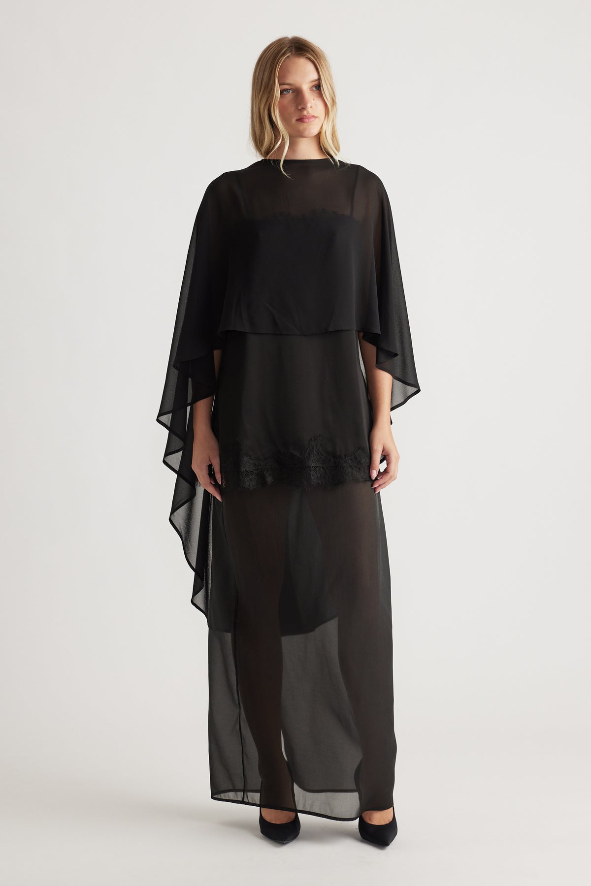 Front view of a black flowing georgette cape top layered over a sheer long skirt with lace detail at hem