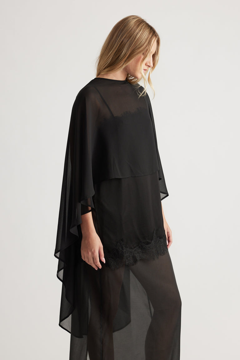 Side view of woman wearing a flowy black georgette cape top with lace trim and sheer layered fabric design