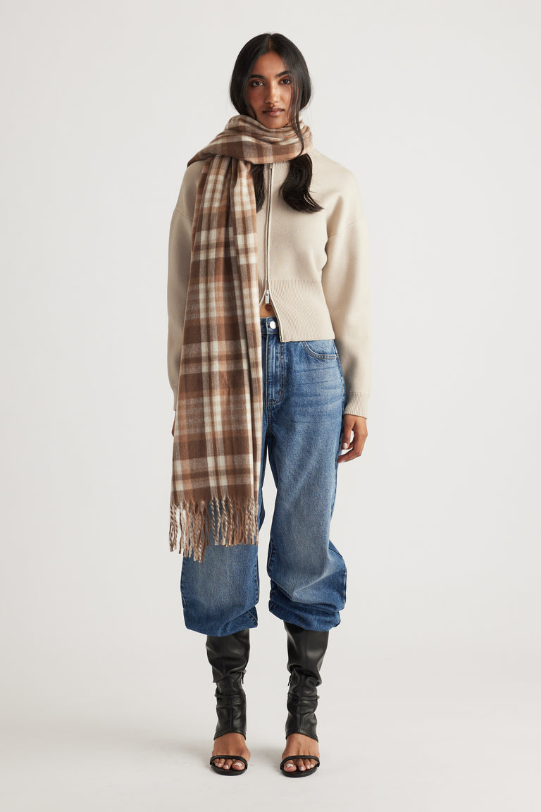 Woman wearing a beige zip-up cardigan with relaxed blue jeans and a long brown check scarf wrapped around her neck