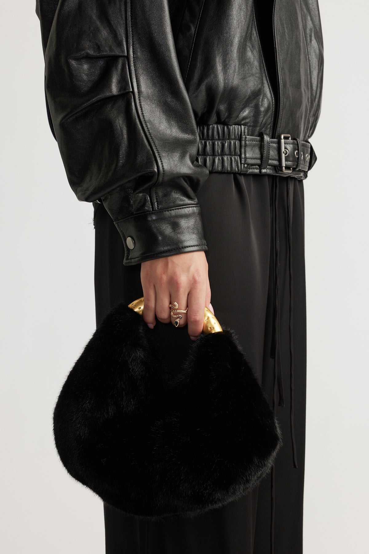 Hand wearing a gold snake-inspired ring holding a black plush bag with a curved gold handle, styled with a black leather jacket.