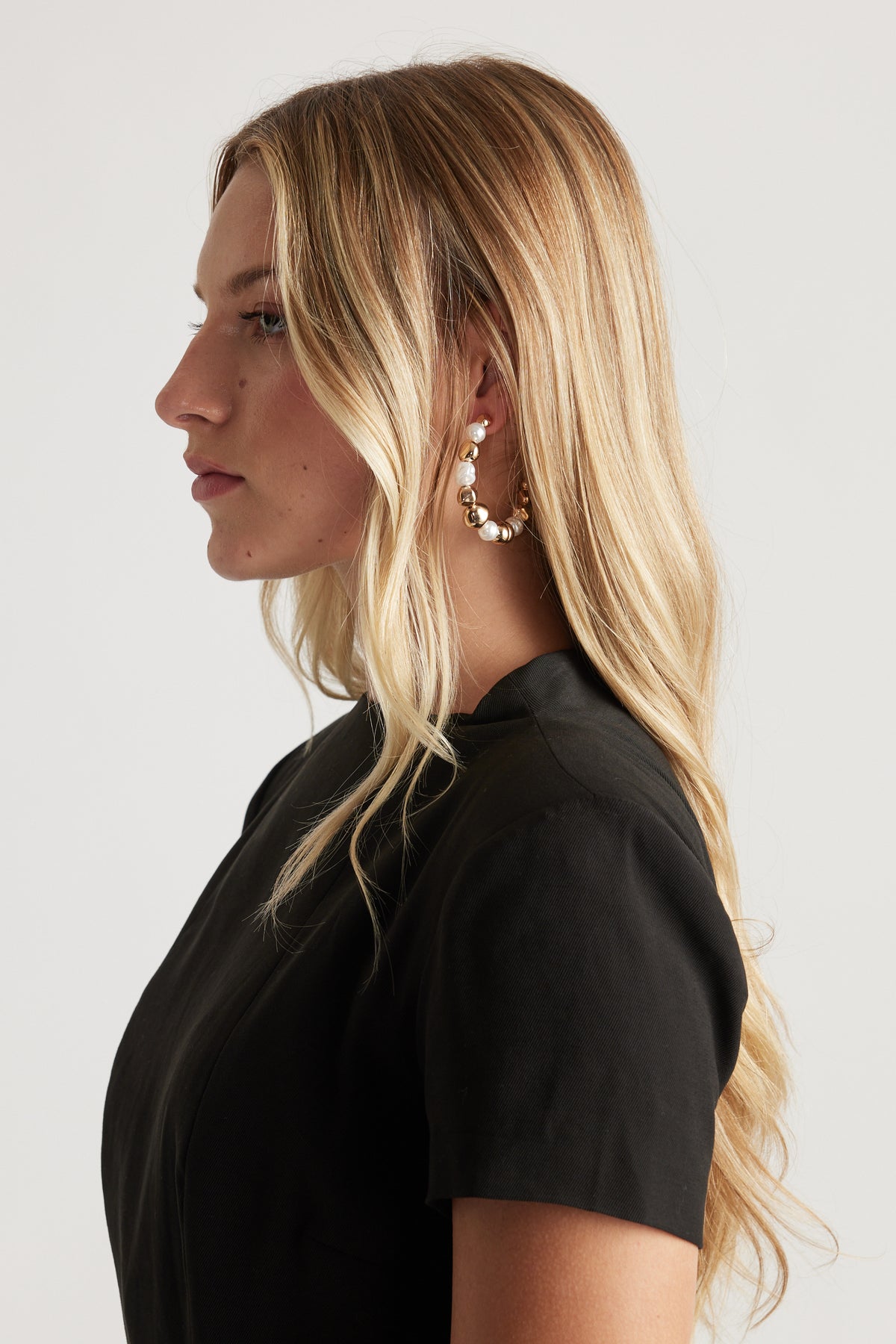 Profile of woman with long blonde hair wearing gold hoop earrings adorned with white pearls and a black short-sleeve top