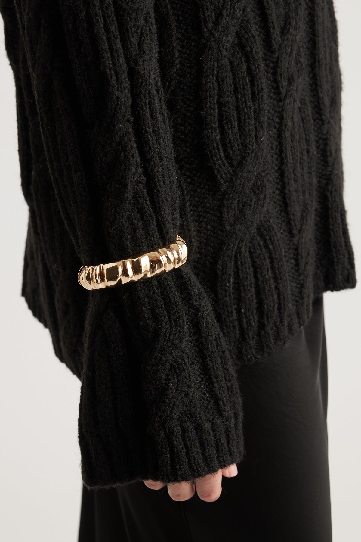 Chunky gold cuff bracelet with a textured design worn over the sleeve of a black knit sweater.