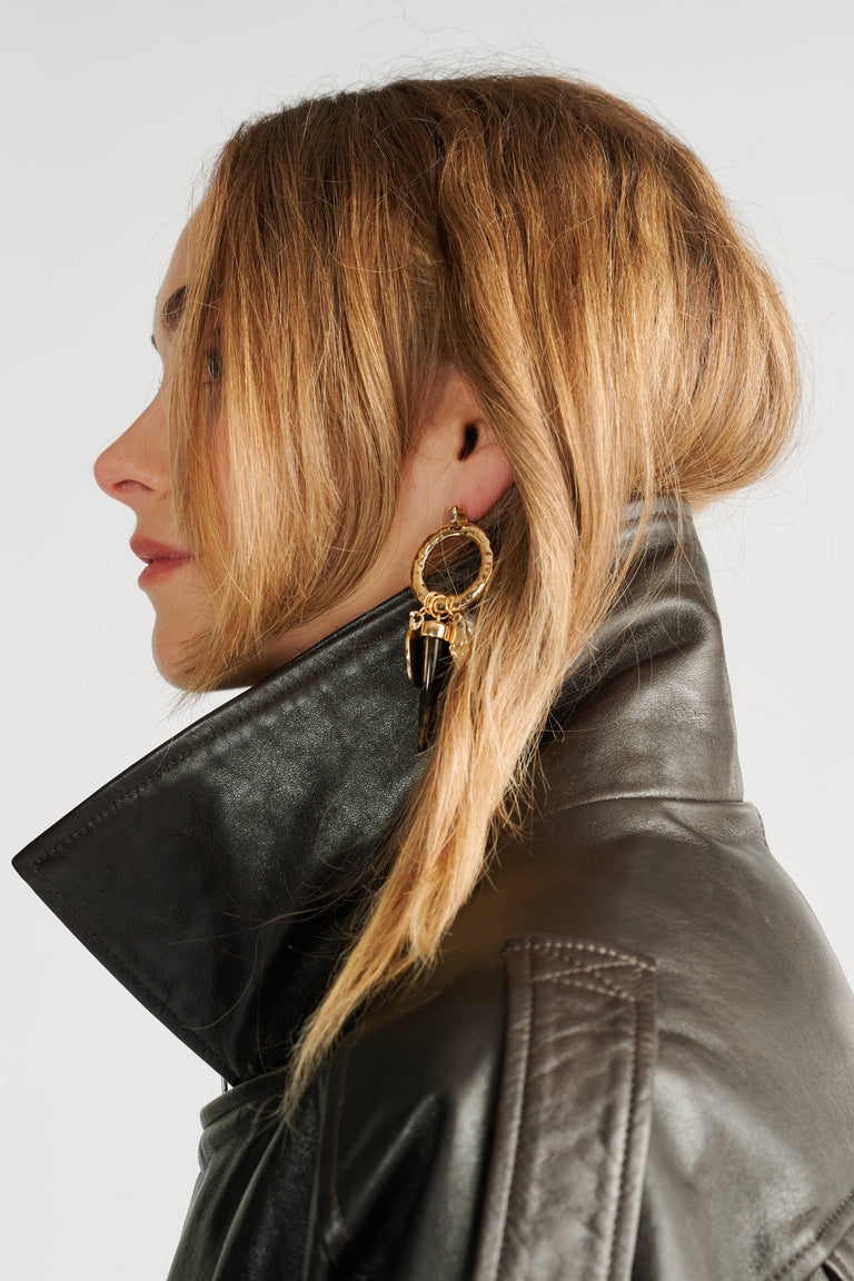 Side profile of a woman wearing textured gold hoop earrings with black charm details and a black leather jacket collar upturned.