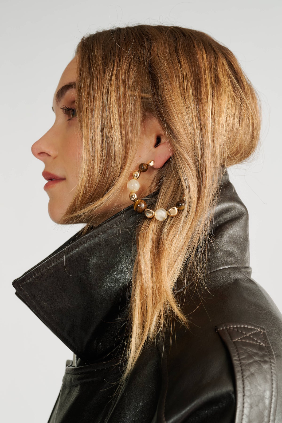 Side profile of woman with wavy blonde hair wearing large gold and stone hoop earrings and a high-collared black leather jacket.