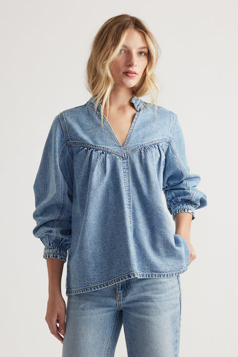 Woman wearing a light blue relaxed denim blouse with gathered detailing and a notched collar paired with blue jeans.