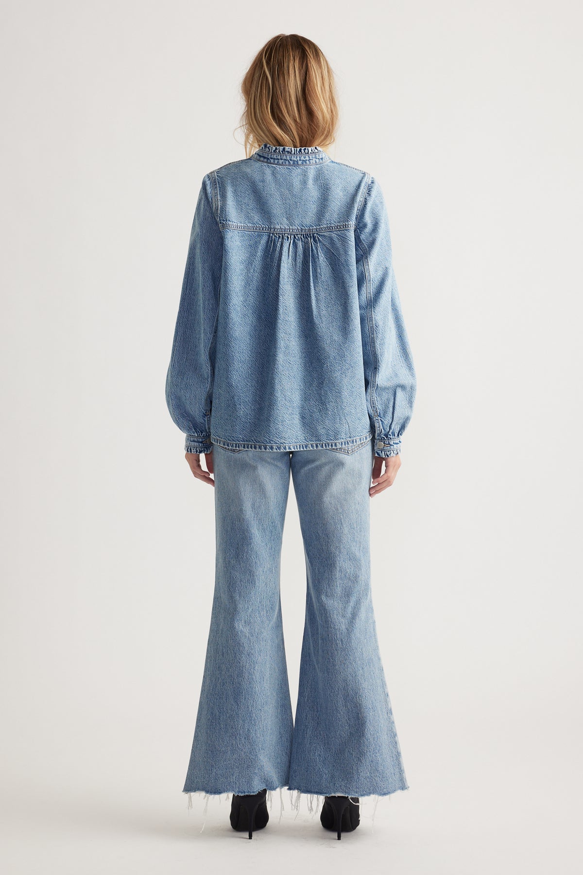 Woman wearing a light blue denim blouse with gathered yoke detail paired with flared raw-hem jeans and black boots