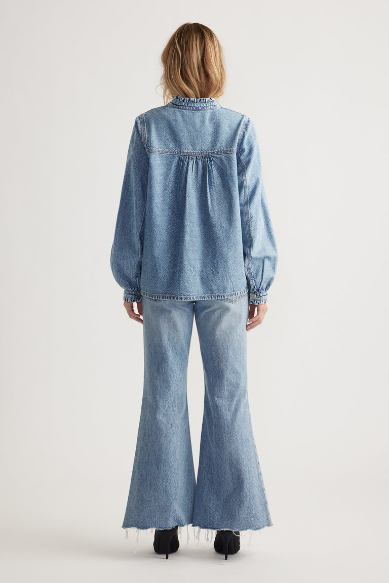 Woman wearing a light blue denim blouse with gathered yoke detail paired with flared raw-hem jeans and black boots