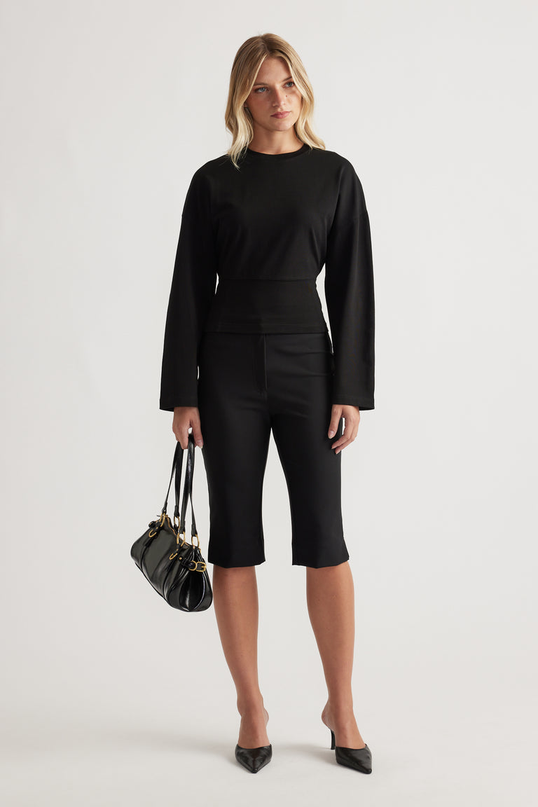 Black long sleeve top with a fitted waist detail, styled with cropped slim black pants and pointed heels.