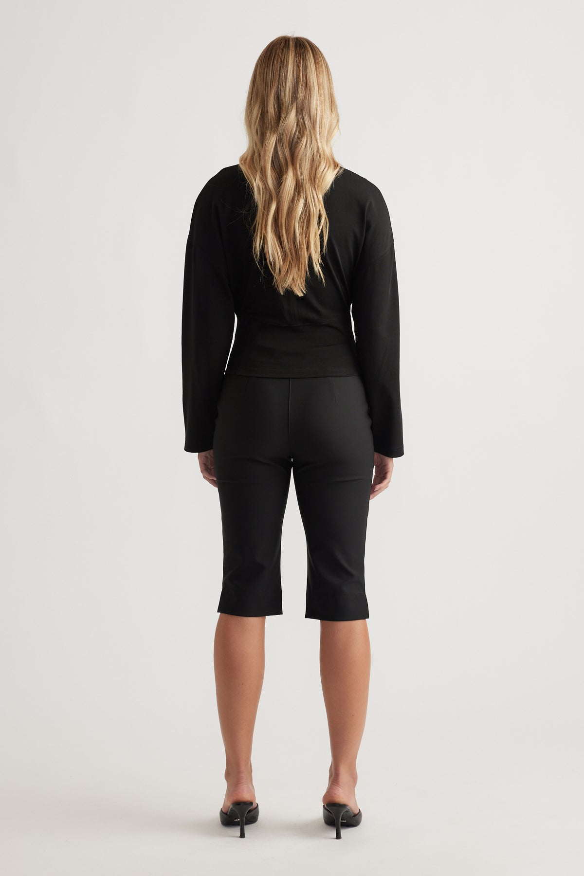 Back view of a fitted black long sleeve top paired with tailored cropped pants and black heeled mules.