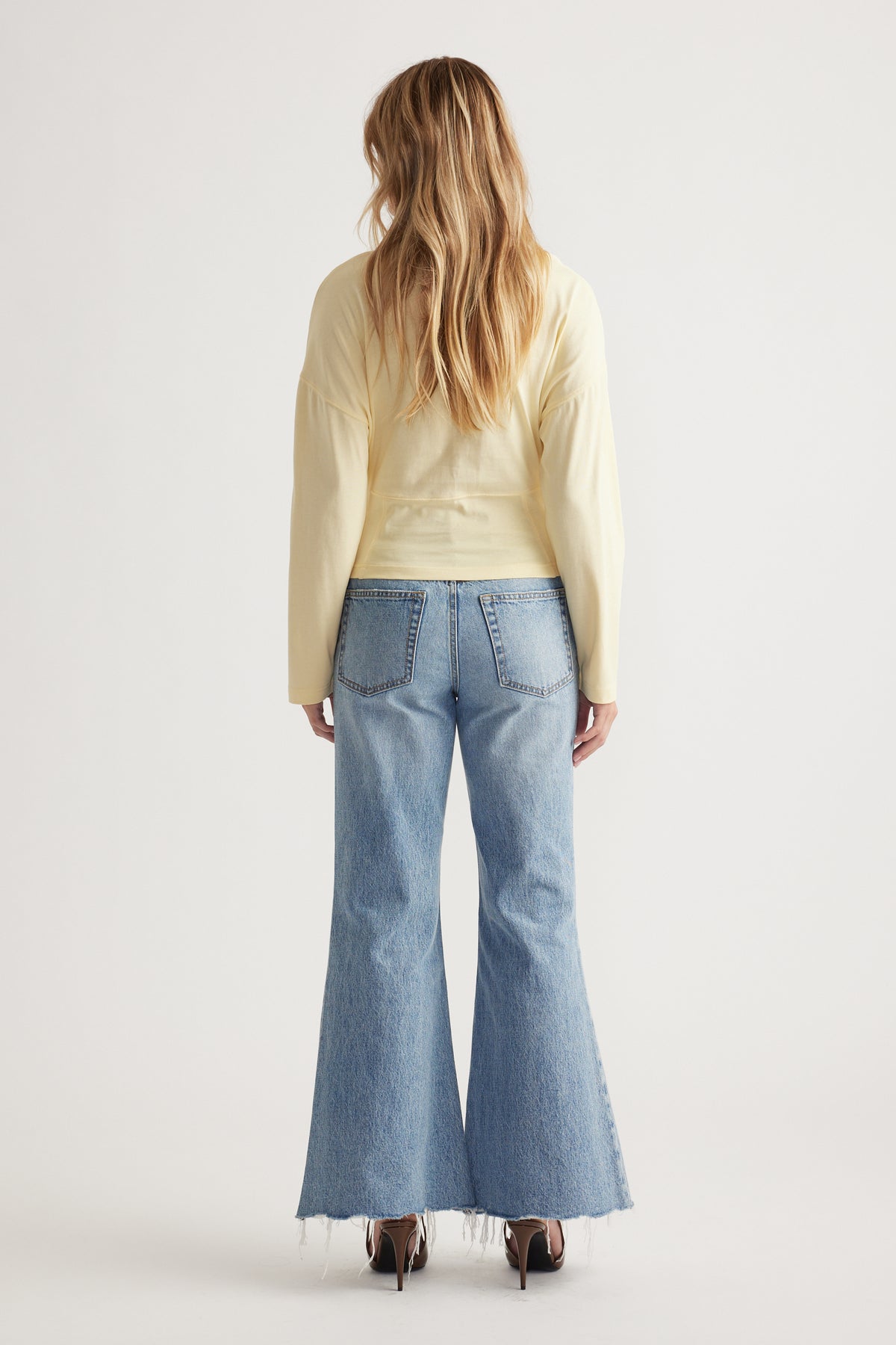 Woman with loose blonde hair wearing a pale yellow long sleeve top paired with light wash flared jeans and heels
