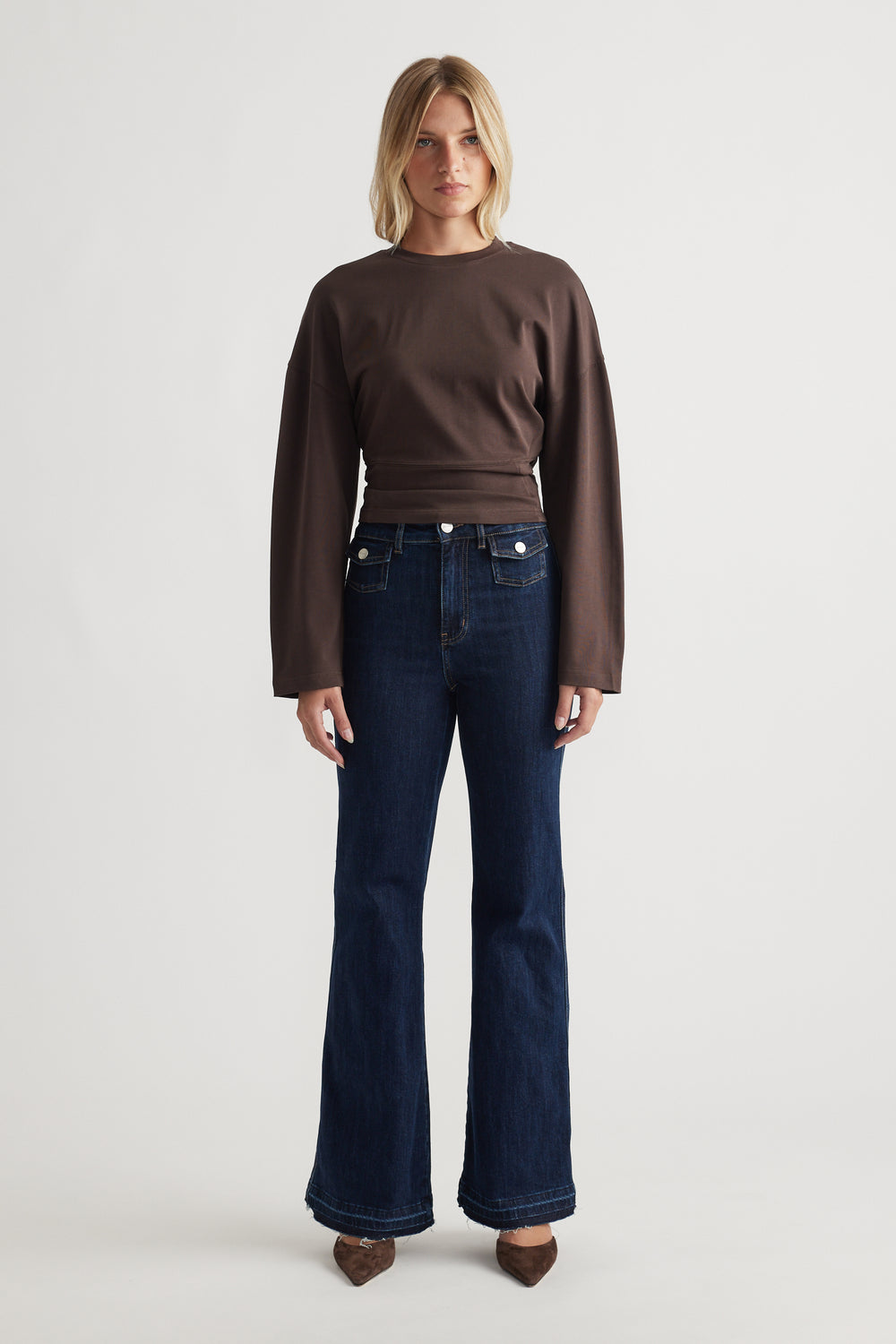 Woman wearing a relaxed fitted long sleeve brown top paired with dark flared denim jeans and pointed brown shoes