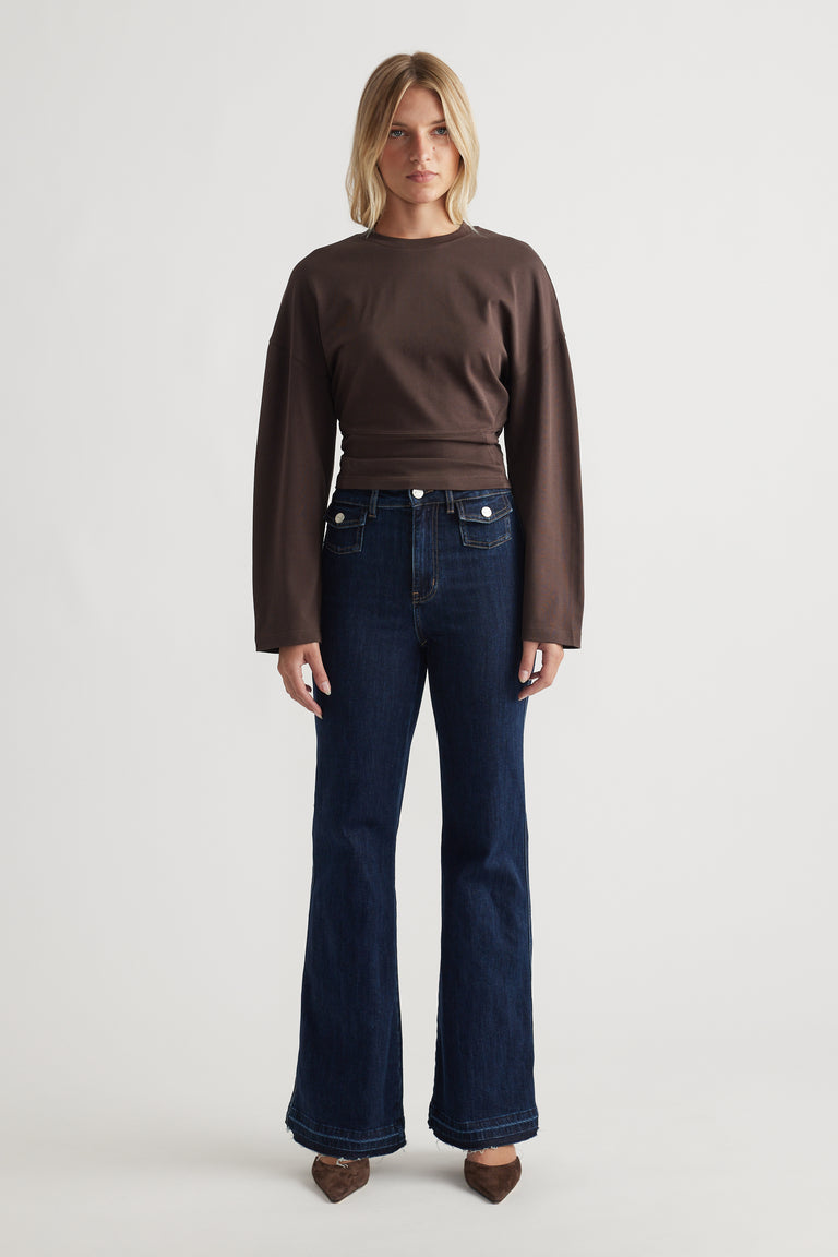 Woman wearing a relaxed fitted long sleeve brown top paired with dark flared denim jeans and pointed brown shoes