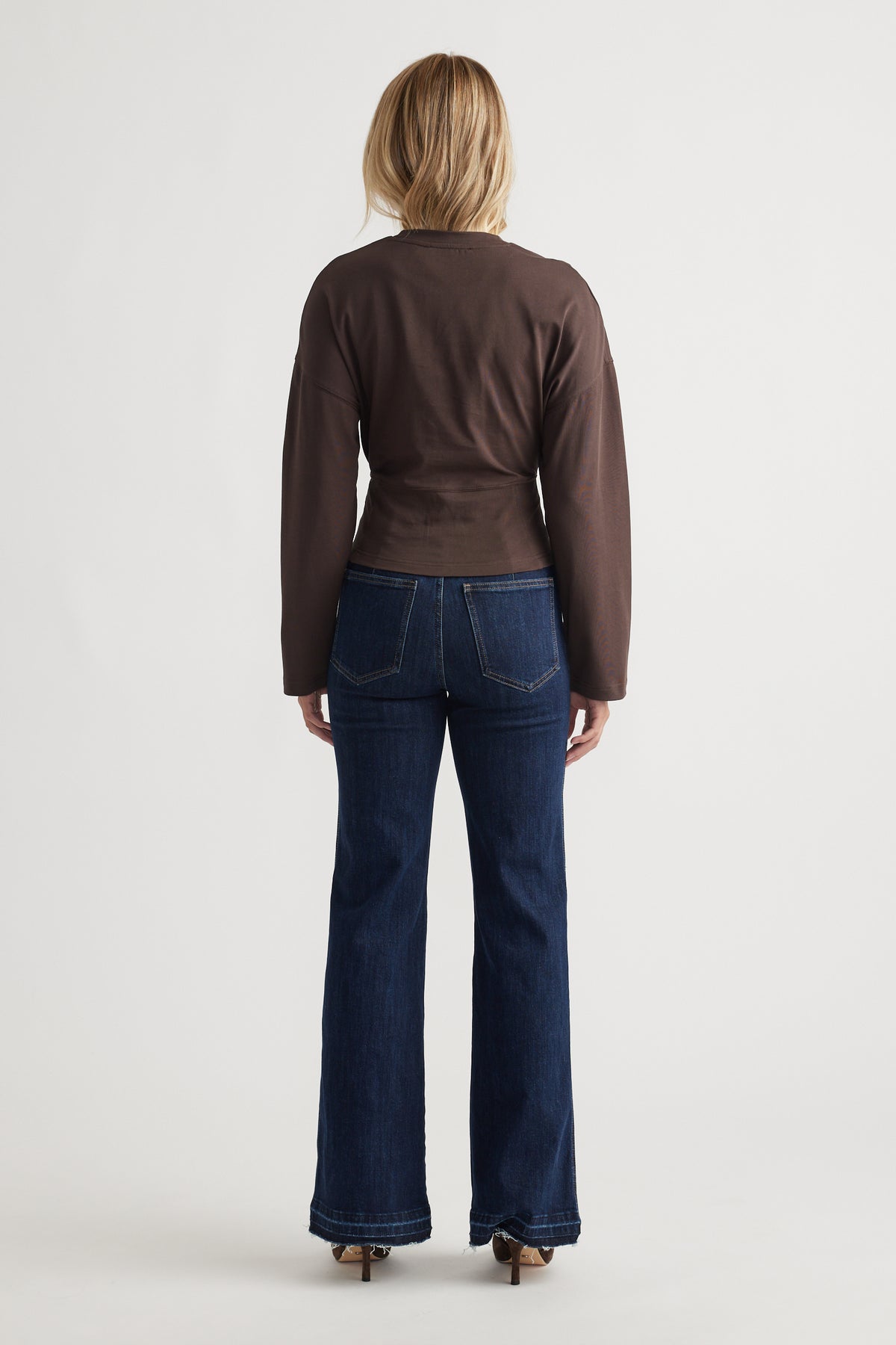 Back view of a fitted brown long sleeve top paired with dark blue bootcut denim jeans and heels for a casual look