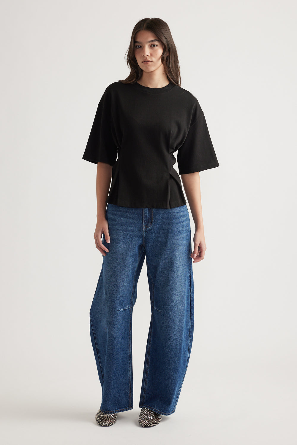 Woman wearing a relaxed black structured tee paired with wide-leg blue jeans and pointed studded shoes against a plain background