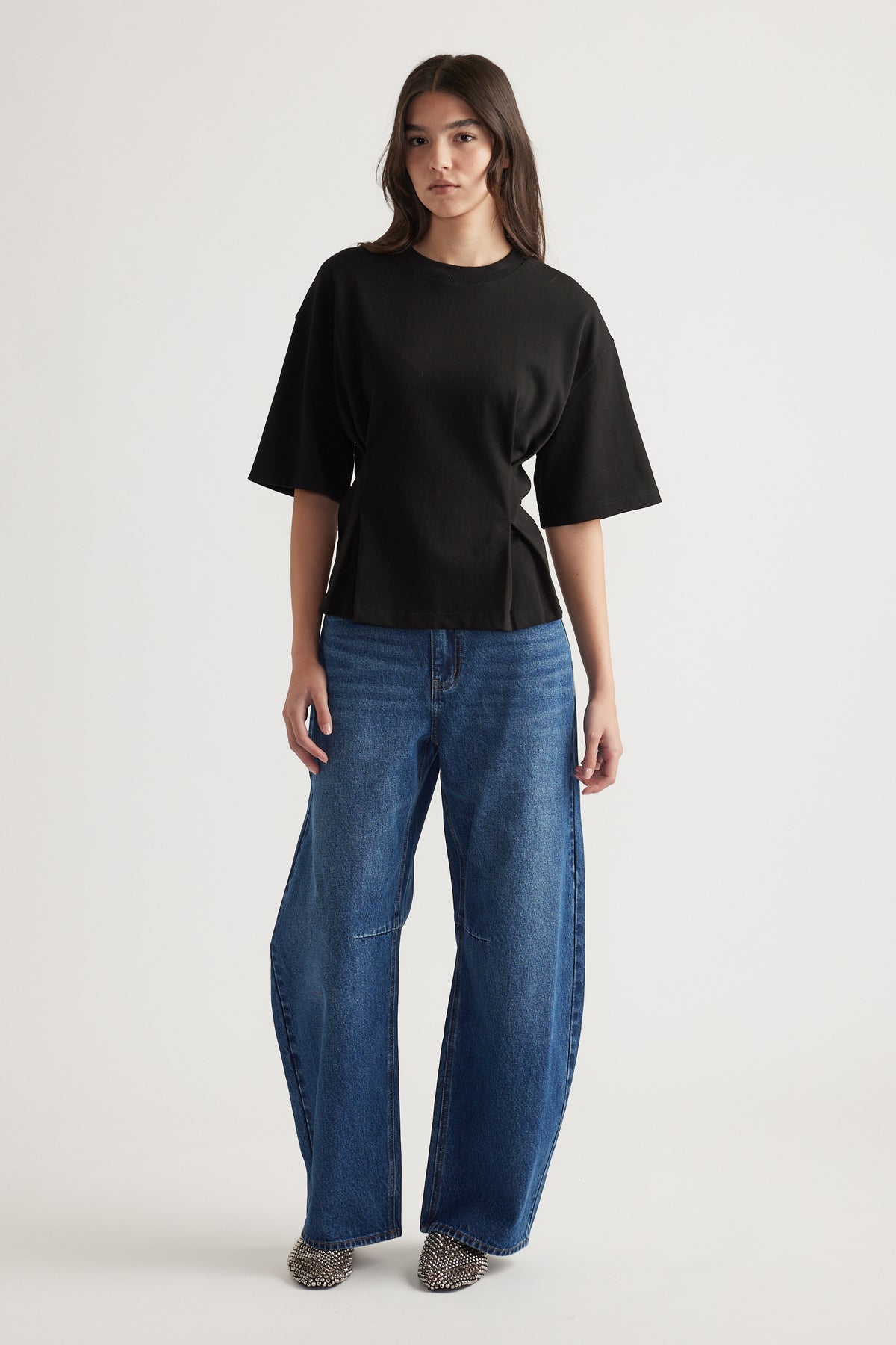Woman wearing a relaxed black structured tee paired with wide-leg blue jeans and pointed studded shoes against a plain background