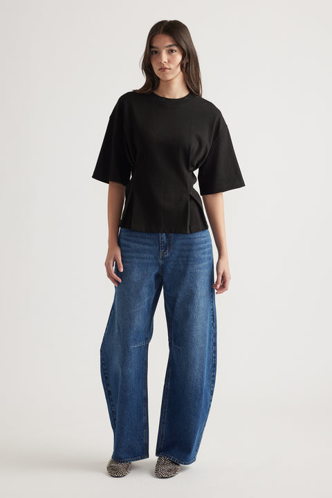 Woman wearing a relaxed black structured tee paired with wide-leg blue jeans and pointed studded shoes against a plain background