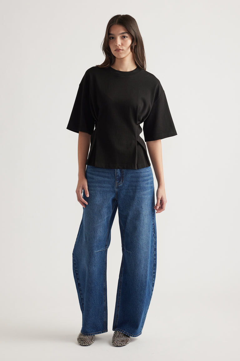 Woman wearing a relaxed black structured tee paired with wide-leg blue jeans and pointed studded shoes against a plain background