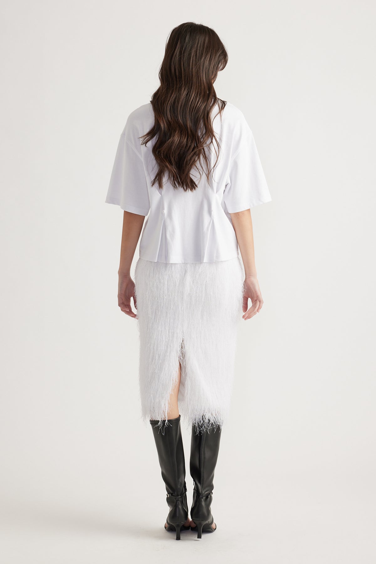 Woman with loose waves wearing a white structured tee paired with a textured white midi skirt and black knee-high boots.