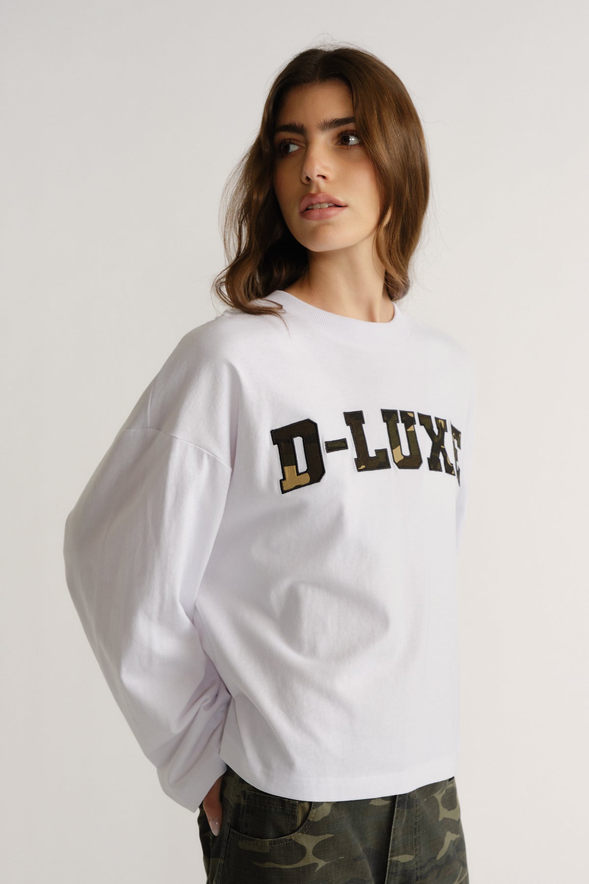 Woman wearing a relaxed white long-sleeve tee with bold "D-Luxe" lettering paired with camo pants in a casual pose