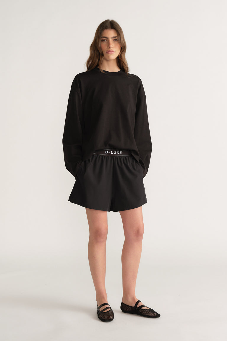 Model wearing an oversized black D LUXE long sleeve tee with black shorts and flat sandals