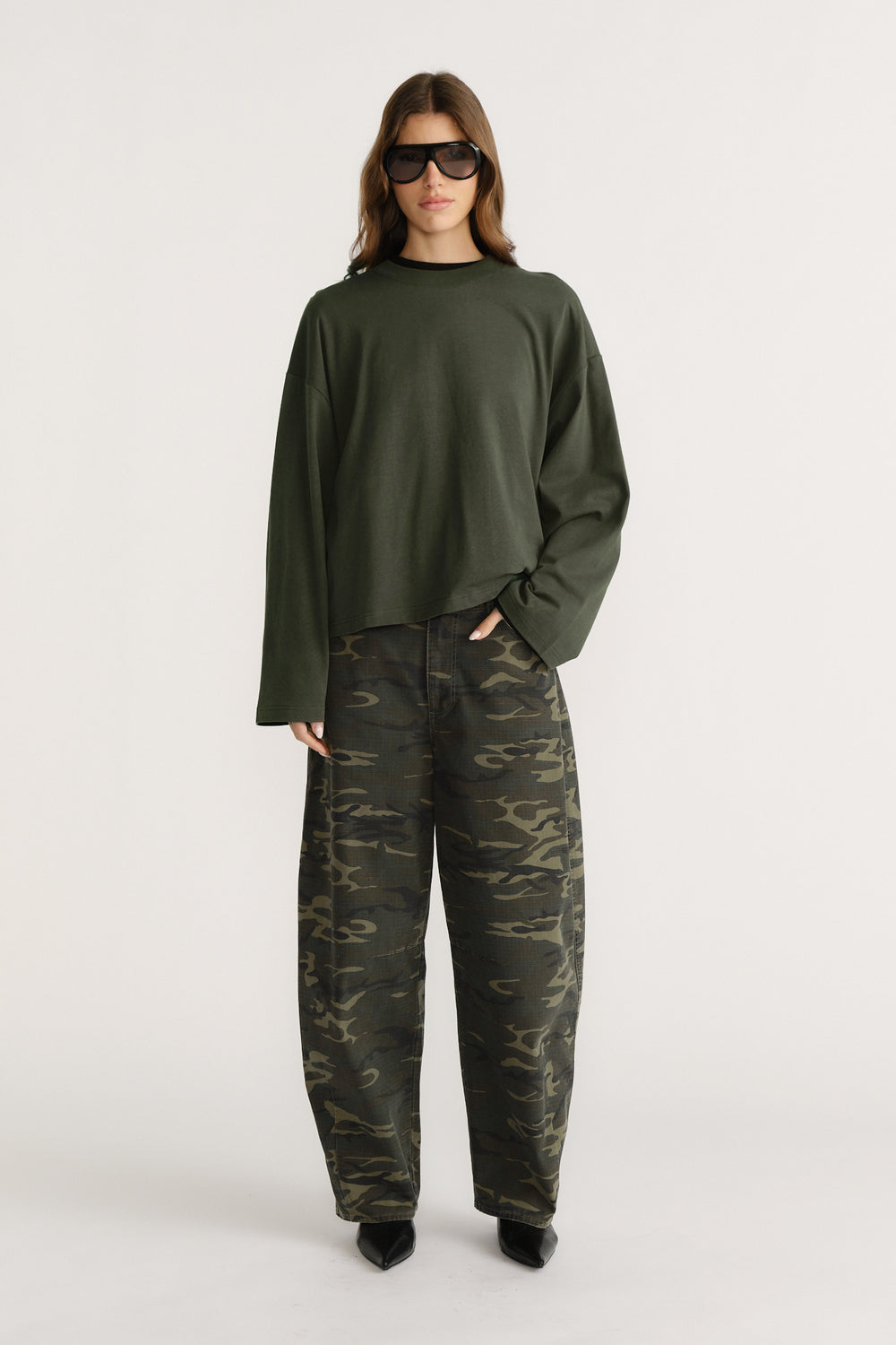 Woman wearing oversized dark green long sleeve tee with wide sleeves paired with loose camo pants and black pointed shoes