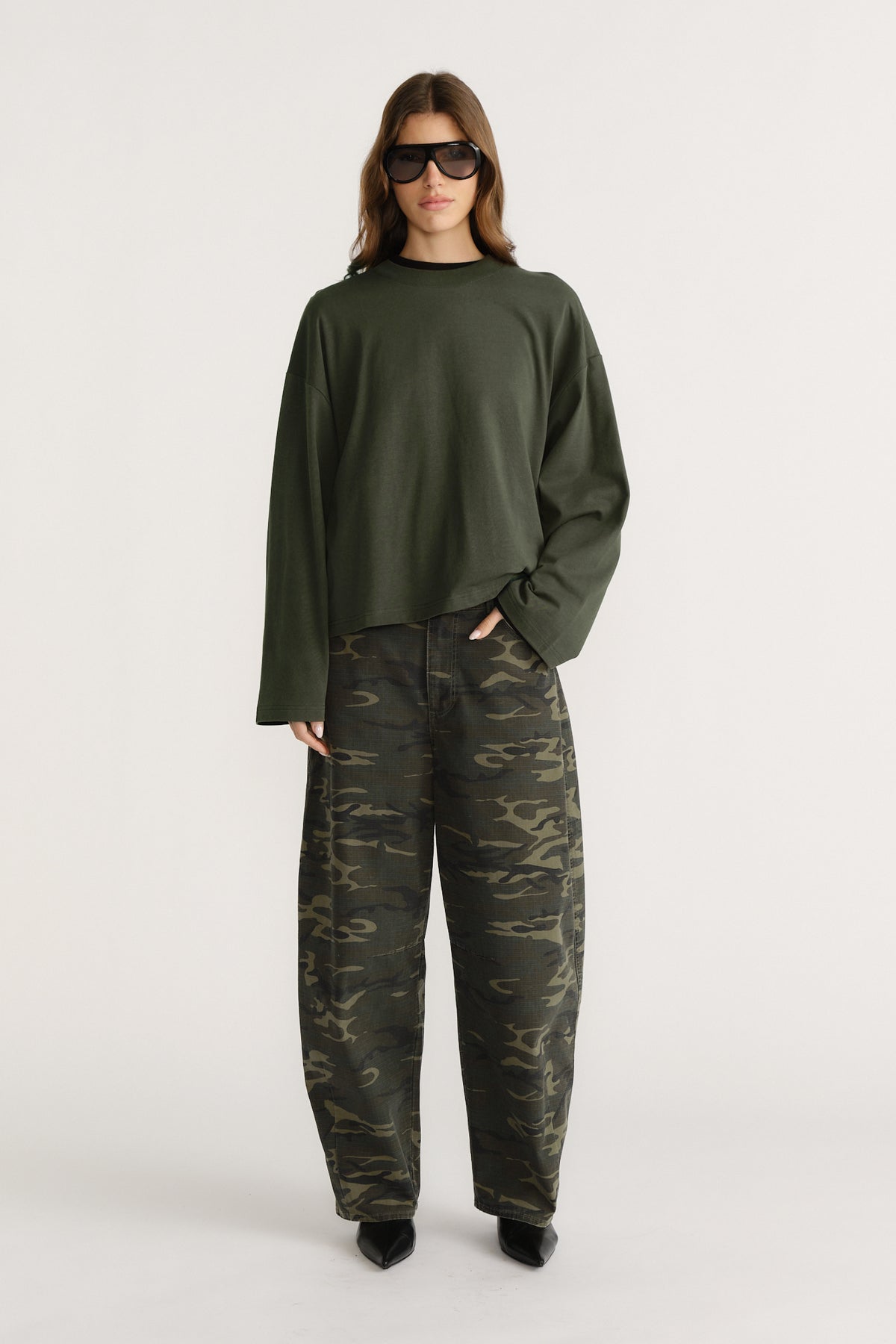 Woman wearing oversized dark green long sleeve tee with wide sleeves paired with loose camo pants and black pointed shoes