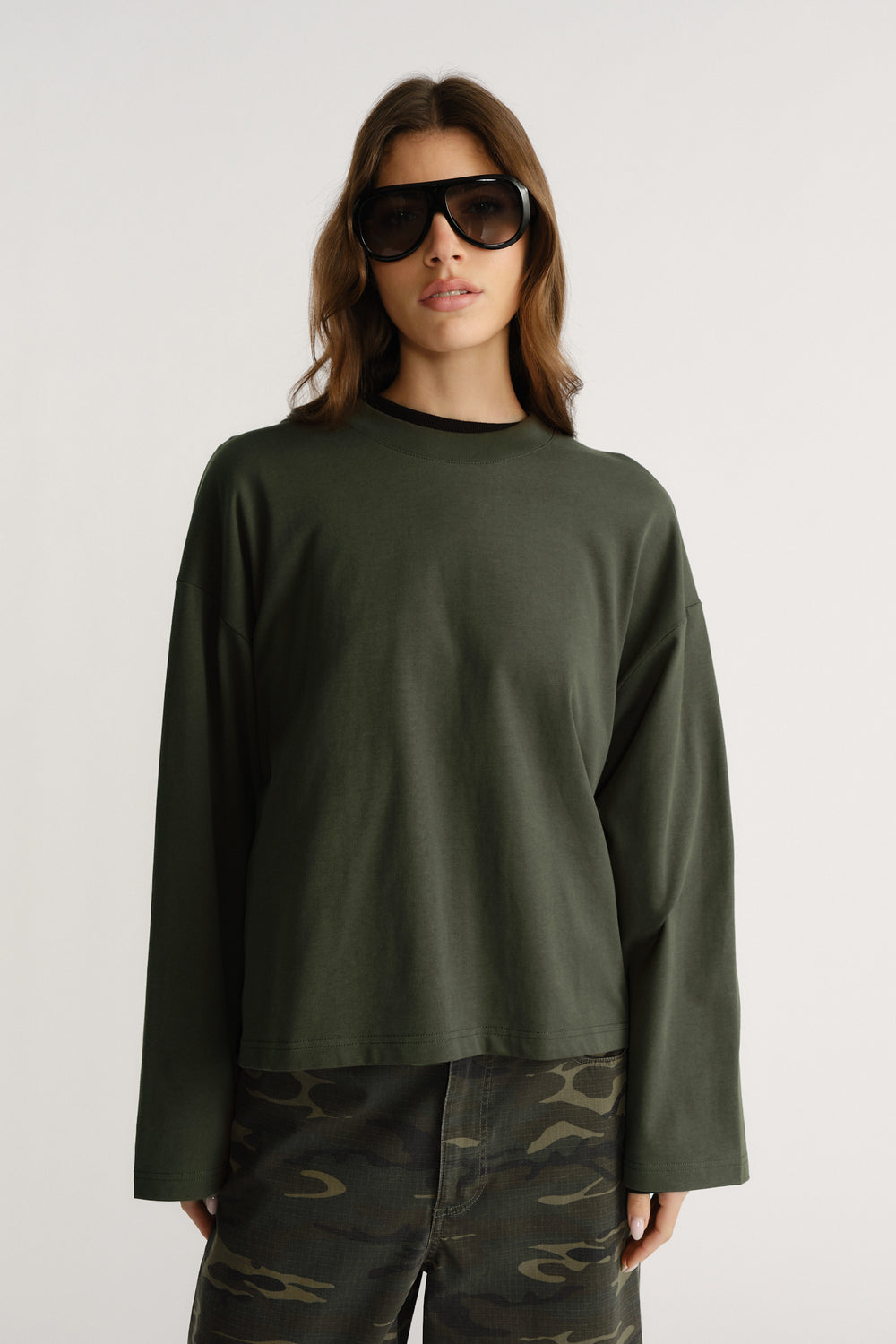Woman wearing boxy dark green long sleeve tee with drop shoulders paired with camo cargo pants and black sunglasses