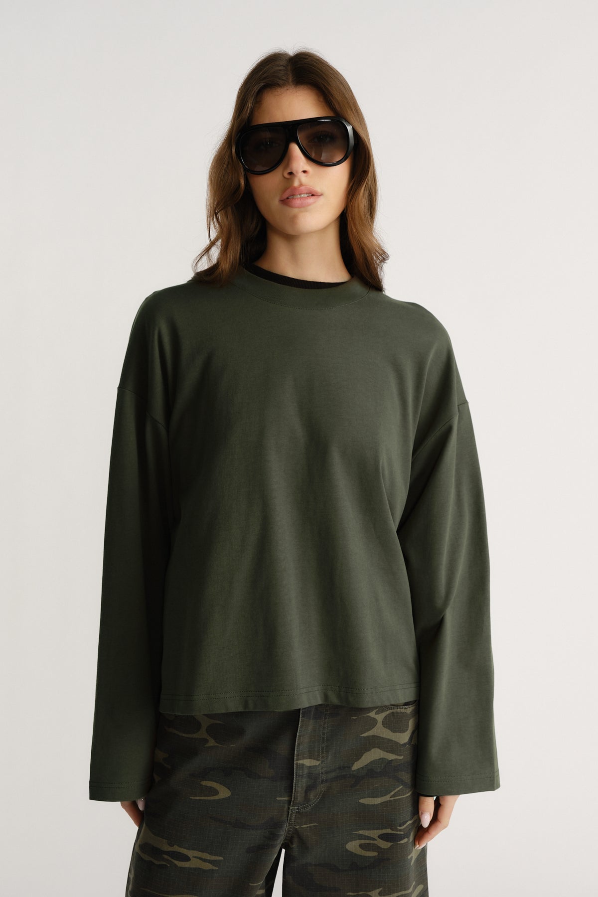Woman wearing boxy dark green long sleeve tee with drop shoulders paired with camo cargo pants and black sunglasses