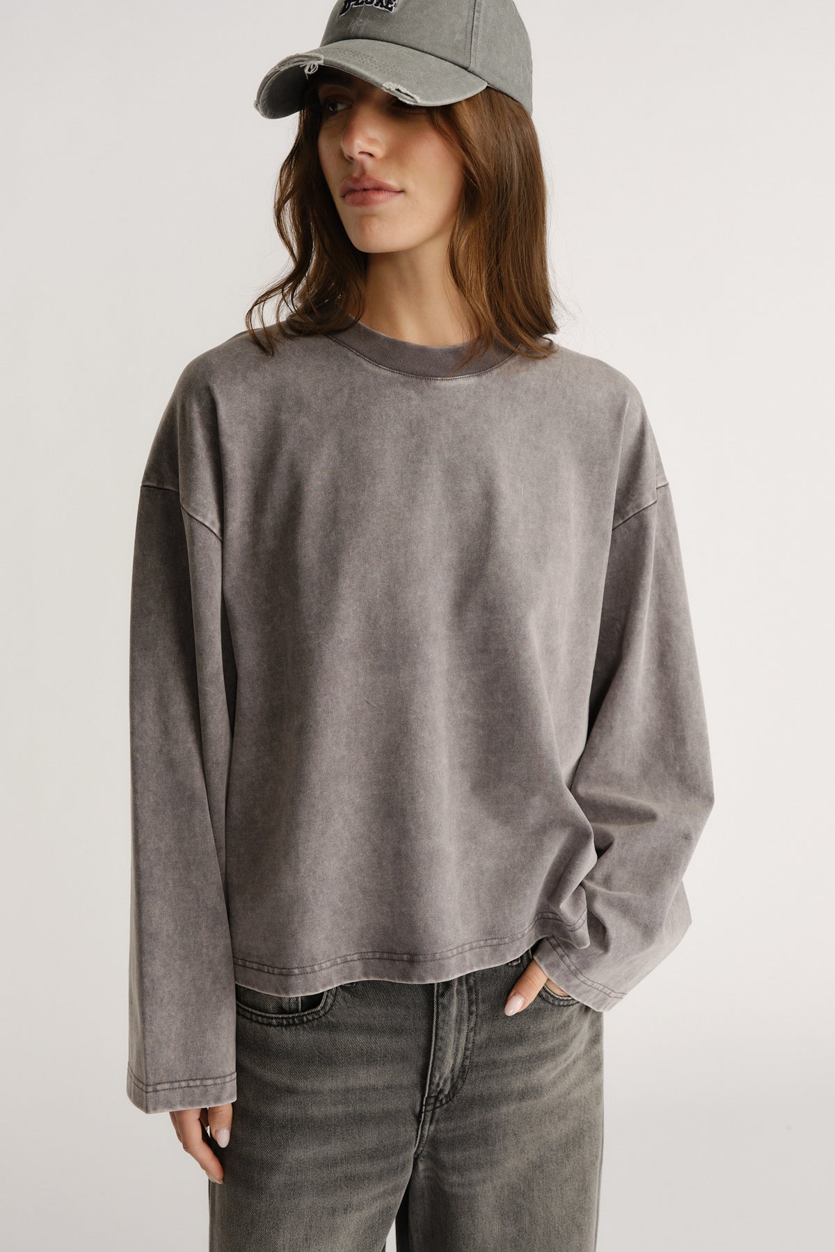 Woman wearing a boxy washed grey long sleeve tee with crew neck and drop shoulders paired with faded grey jeans and a cap