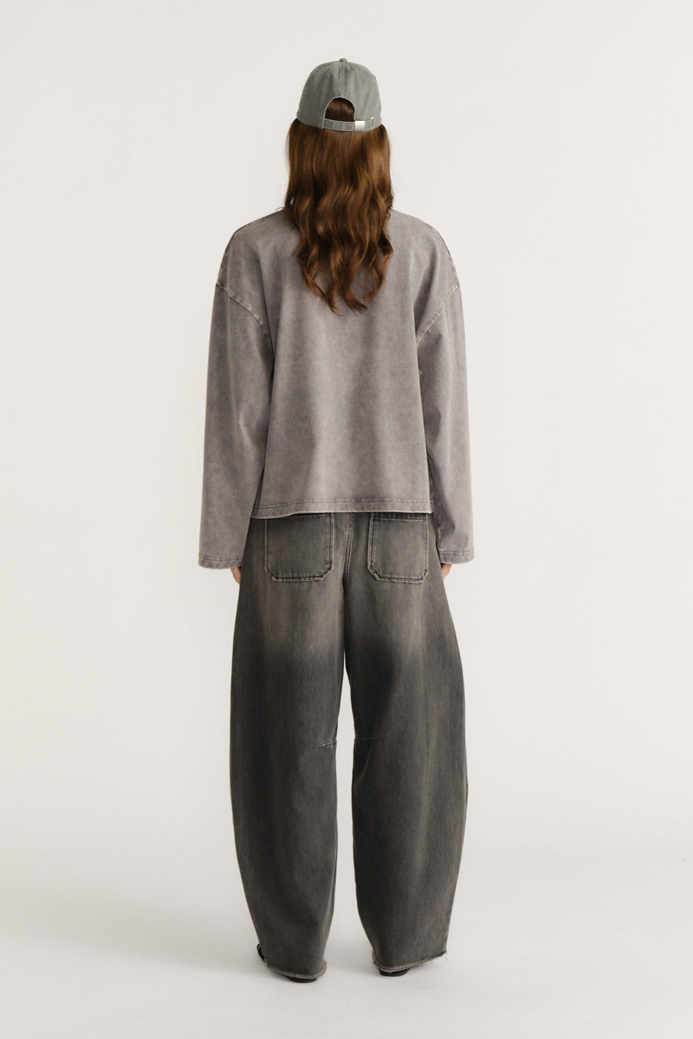 Woman seen from behind wearing a washed grey oversized long sleeve top with wide-leg faded black denim jeans and a grey cap