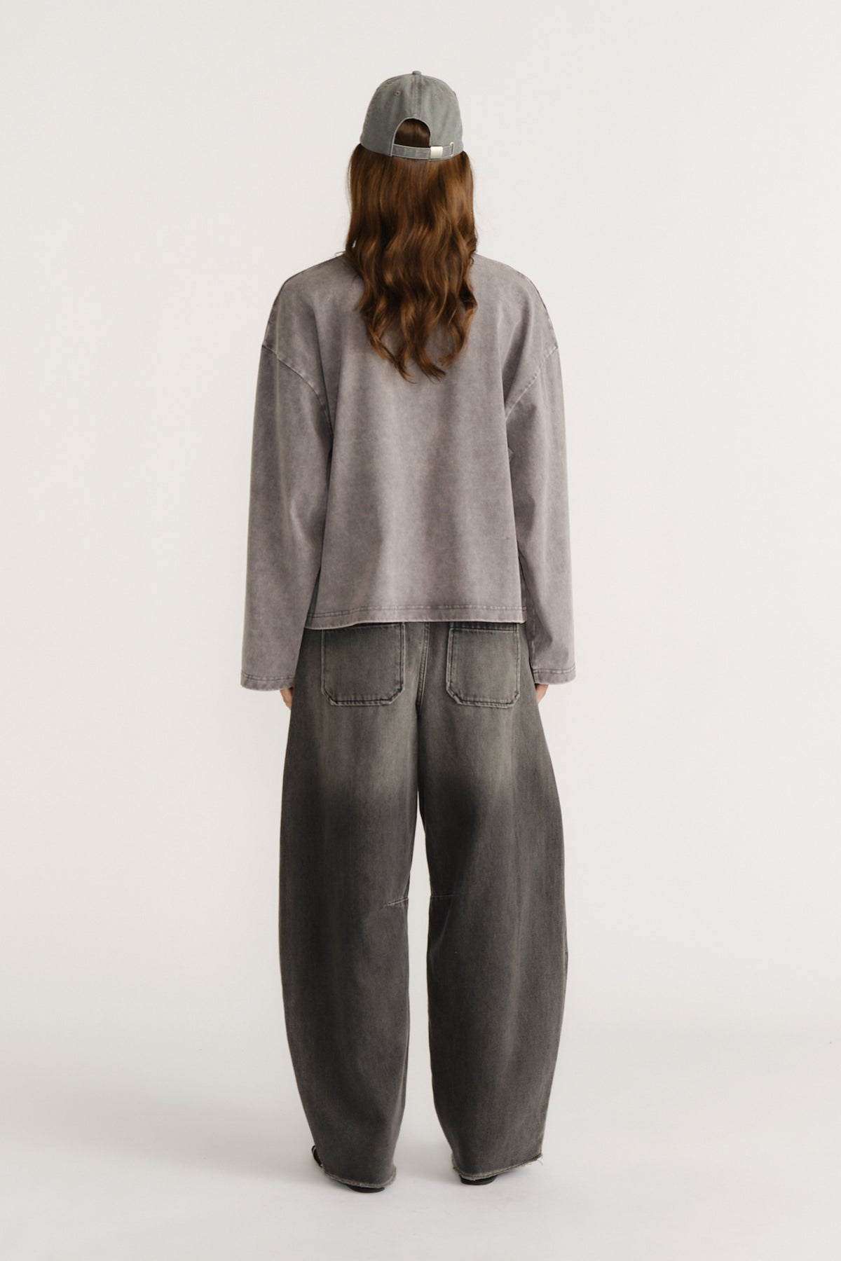 Woman seen from behind wearing a washed grey oversized long sleeve top with wide-leg faded black denim jeans and a grey cap
