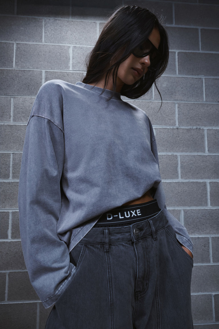 Woman in an oversized grey long sleeve tee and dark jeans, wearing sunglasses