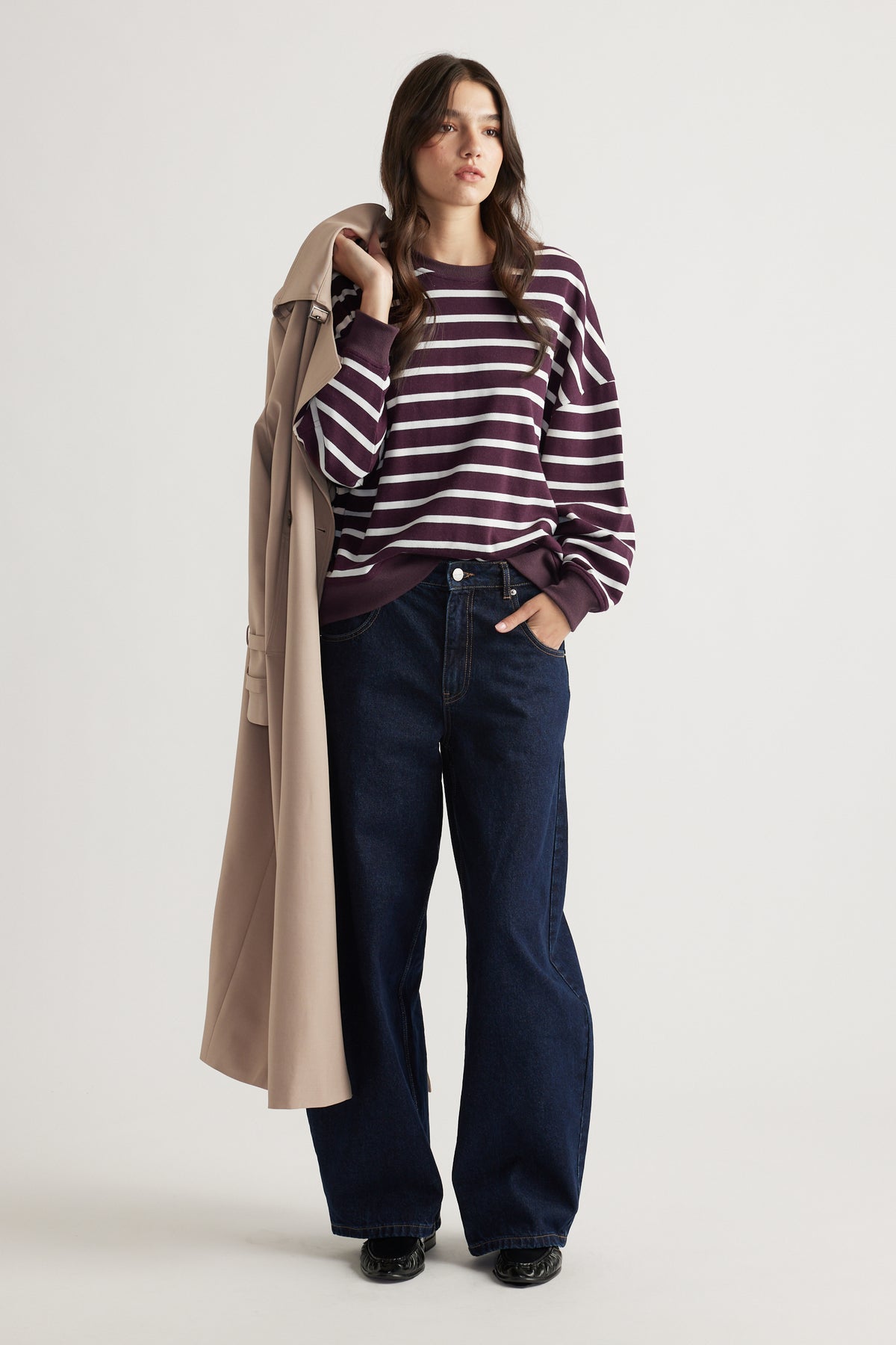 Woman wearing a relaxed striped sweatshirt paired with wide-leg dark denim jeans and holding a beige trench coat over her shoulder