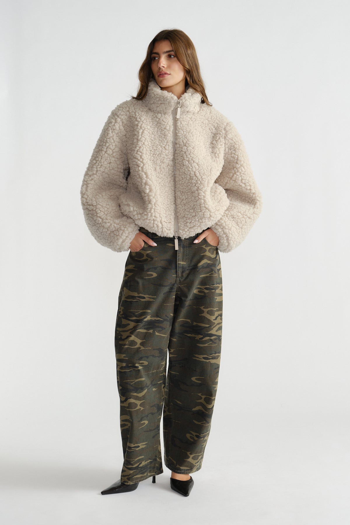 Woman wearing a cream sherpa bomber jacket with high collar and zip front paired with relaxed camo print pants and black heels