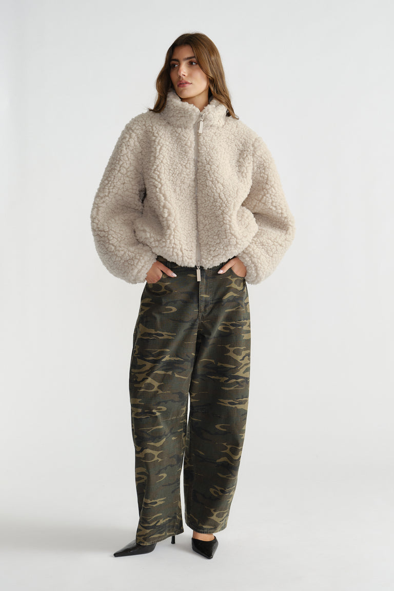 Woman wearing a cream sherpa bomber jacket with high collar and zip front paired with relaxed camo print pants and black heels