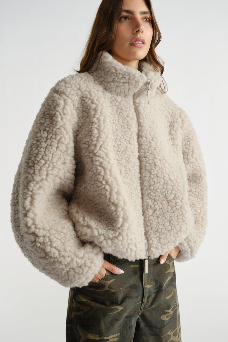 Woman wearing a cream sherpa bomber jacket with a zip front and high collar paired with camo pants