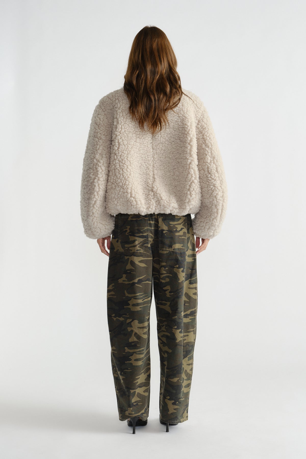 Rear view of woman wearing a soft cream sherpa bomber jacket paired with relaxed-fit camouflage pants and black heels